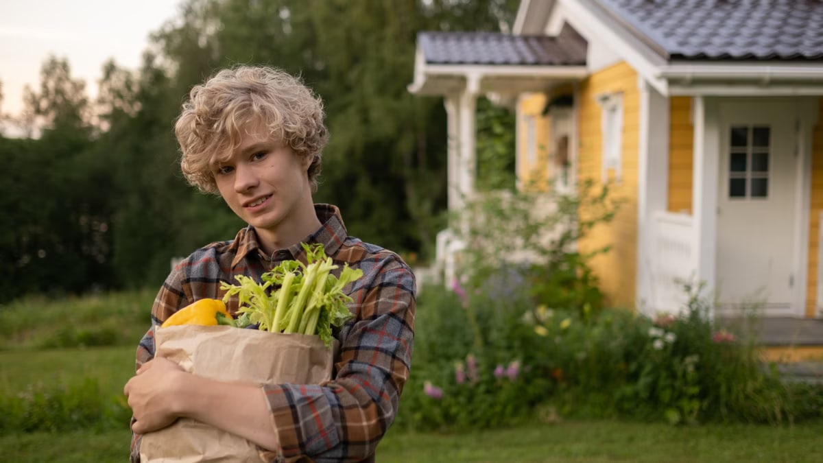Teenage boy holding groceries in a rustic backyard near a charming yellow house.